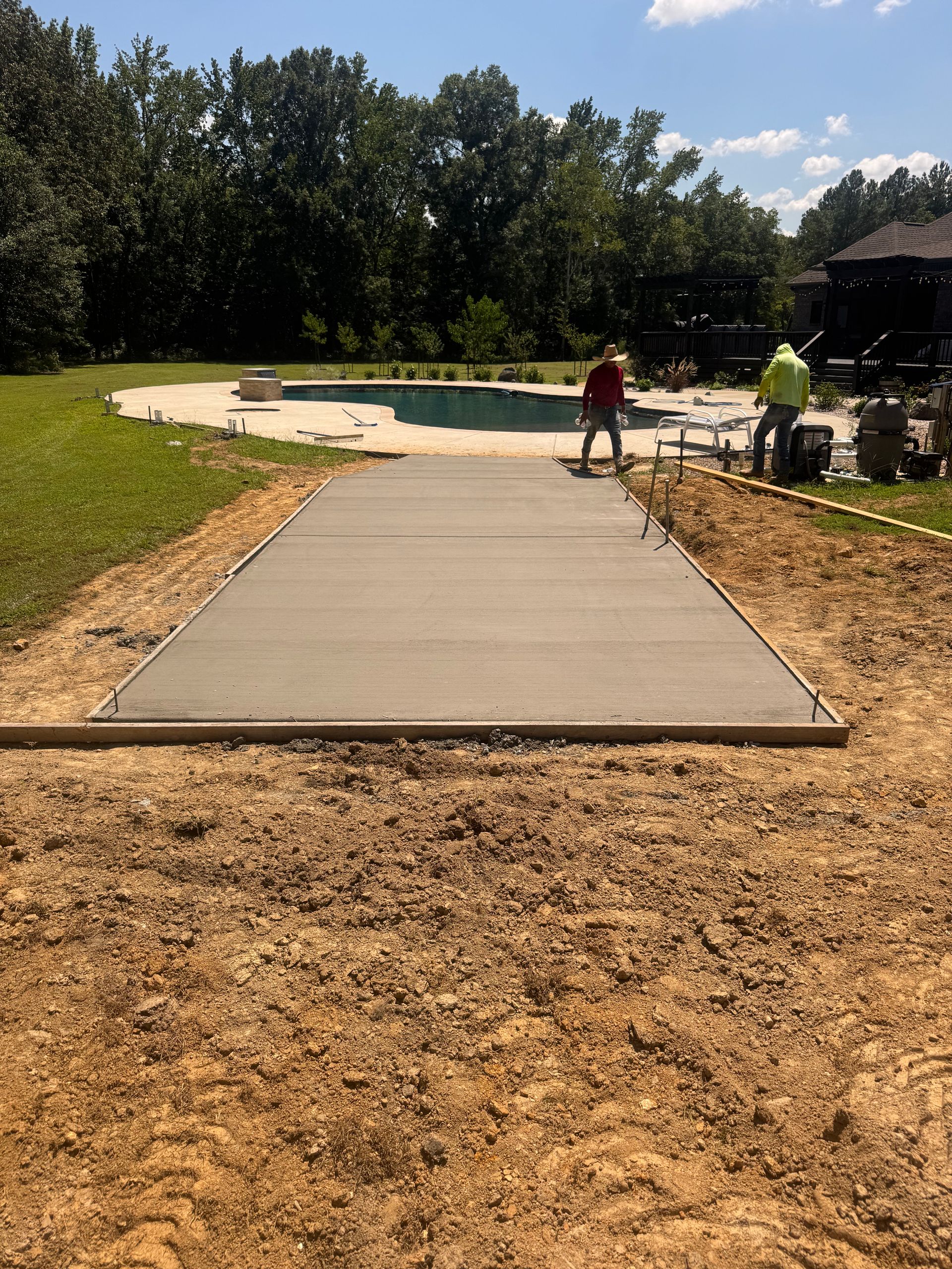 Concrete walkway under construction in a sunny park, with workers and materials in the background