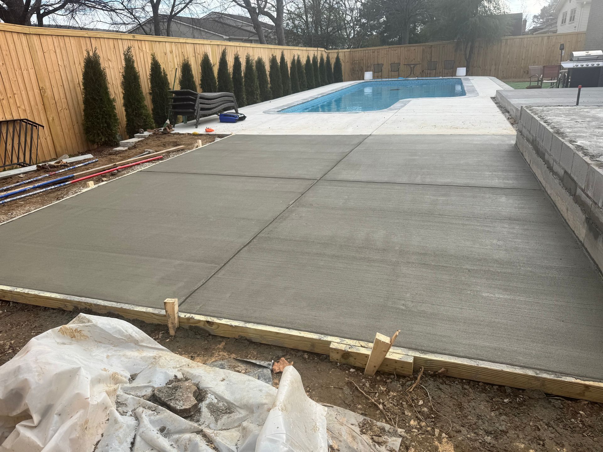 Backyard swimming pool with concrete deck under construction, bordered by wood fence and shrubs