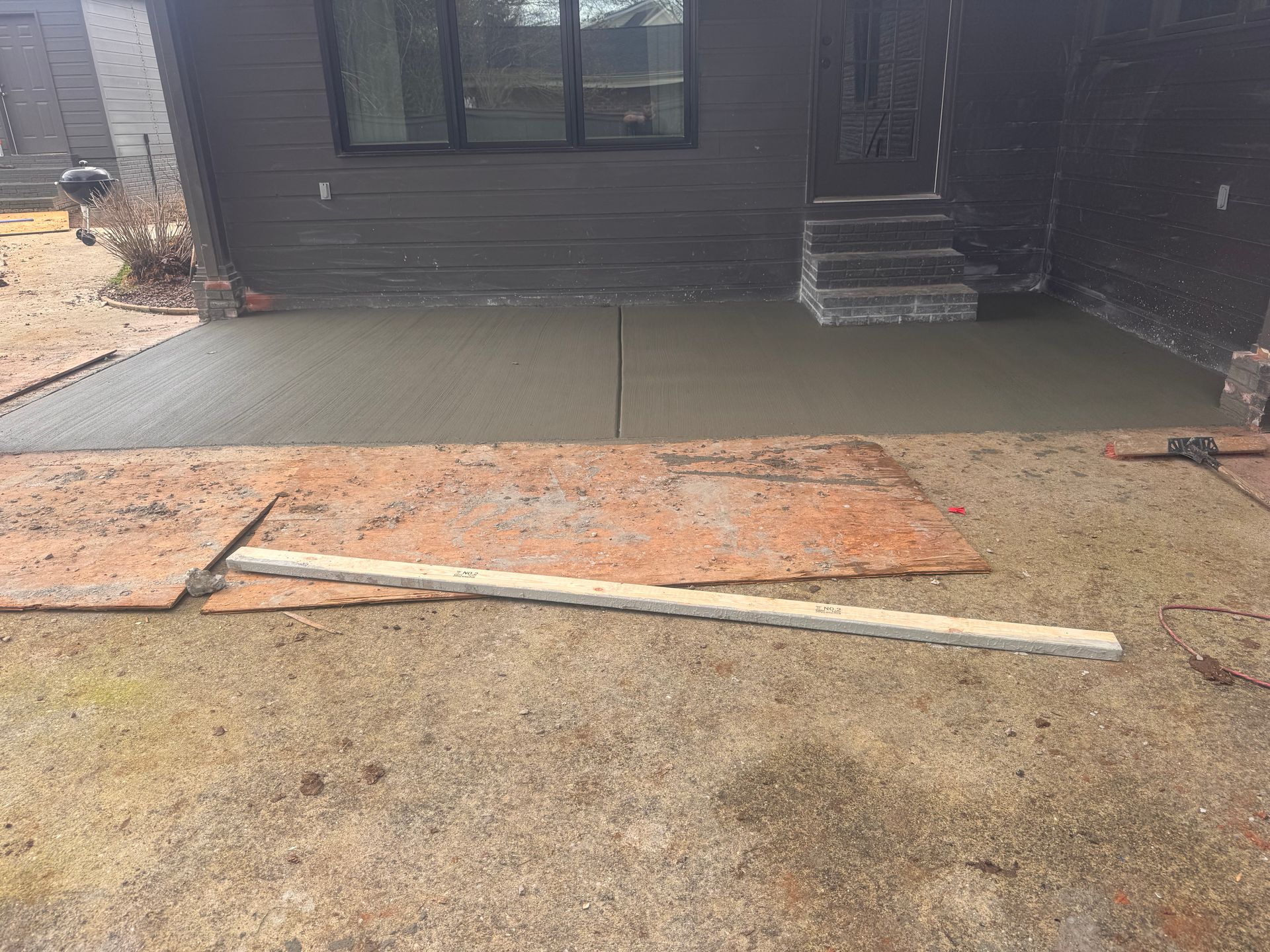Wet concrete patio beside a dark house, with a rusty metal sheet and long board on the ground.
