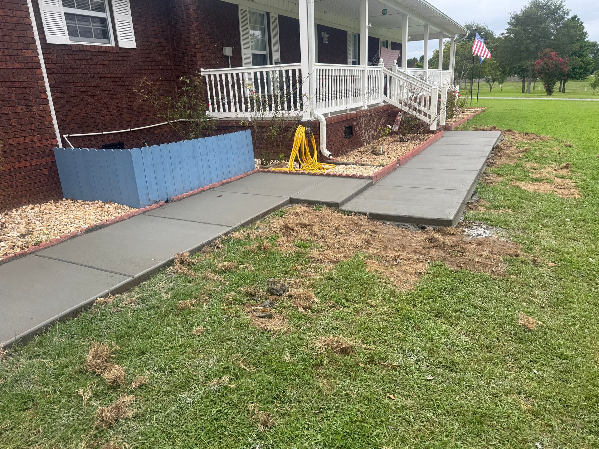 Concrete sidewalk beside a white porch and blue skirting, with a yellow caution sign and patchy lawn.