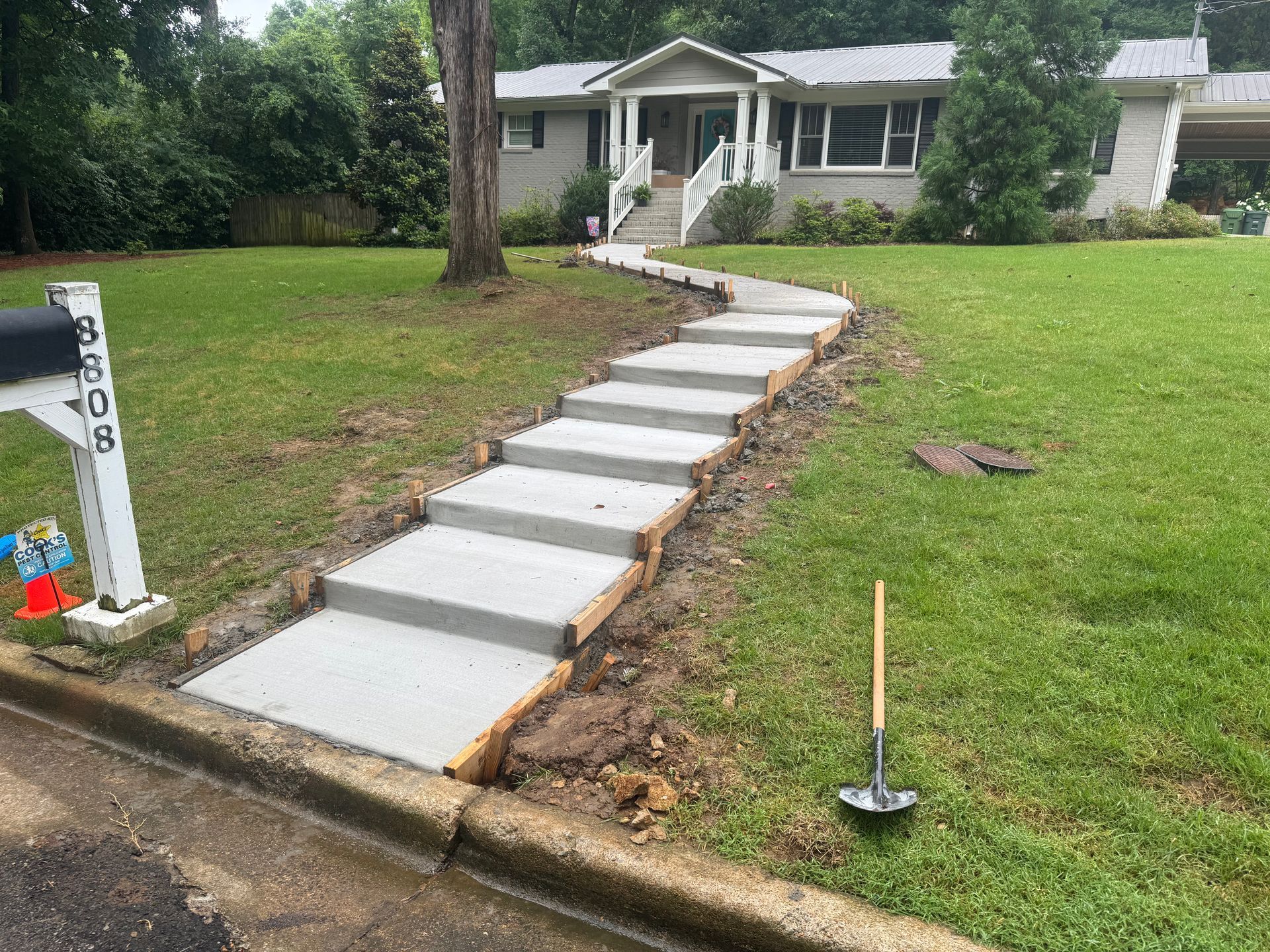 Concrete walkway under construction leading from curb to a house, with a measuring rod and shovel nearby