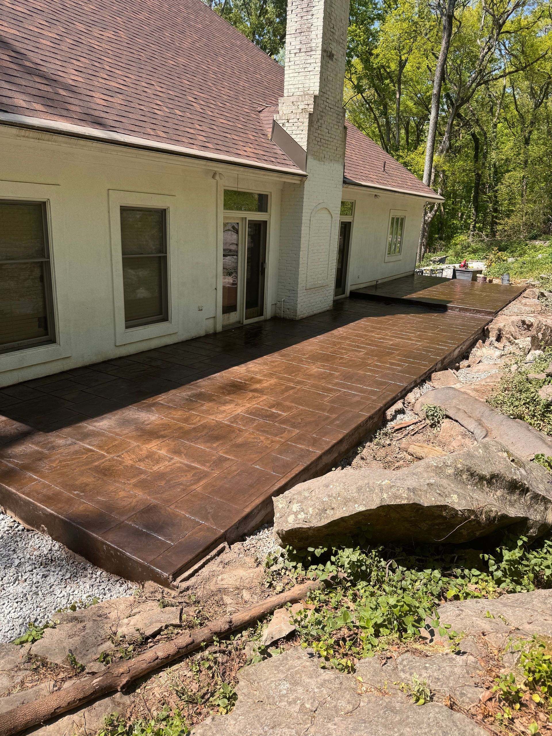 Back patio with brown tile decking beside a white house, bordered by rocks and trees