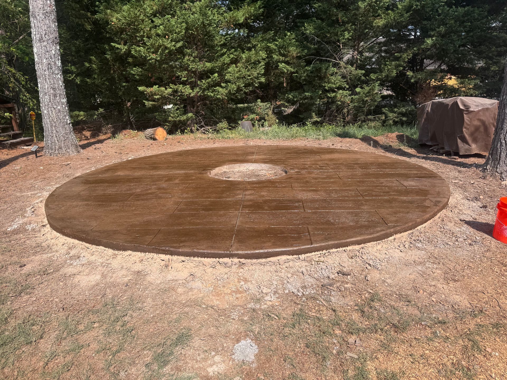 Large circular brown mat or cover laid on bare ground in a wooded backyard area