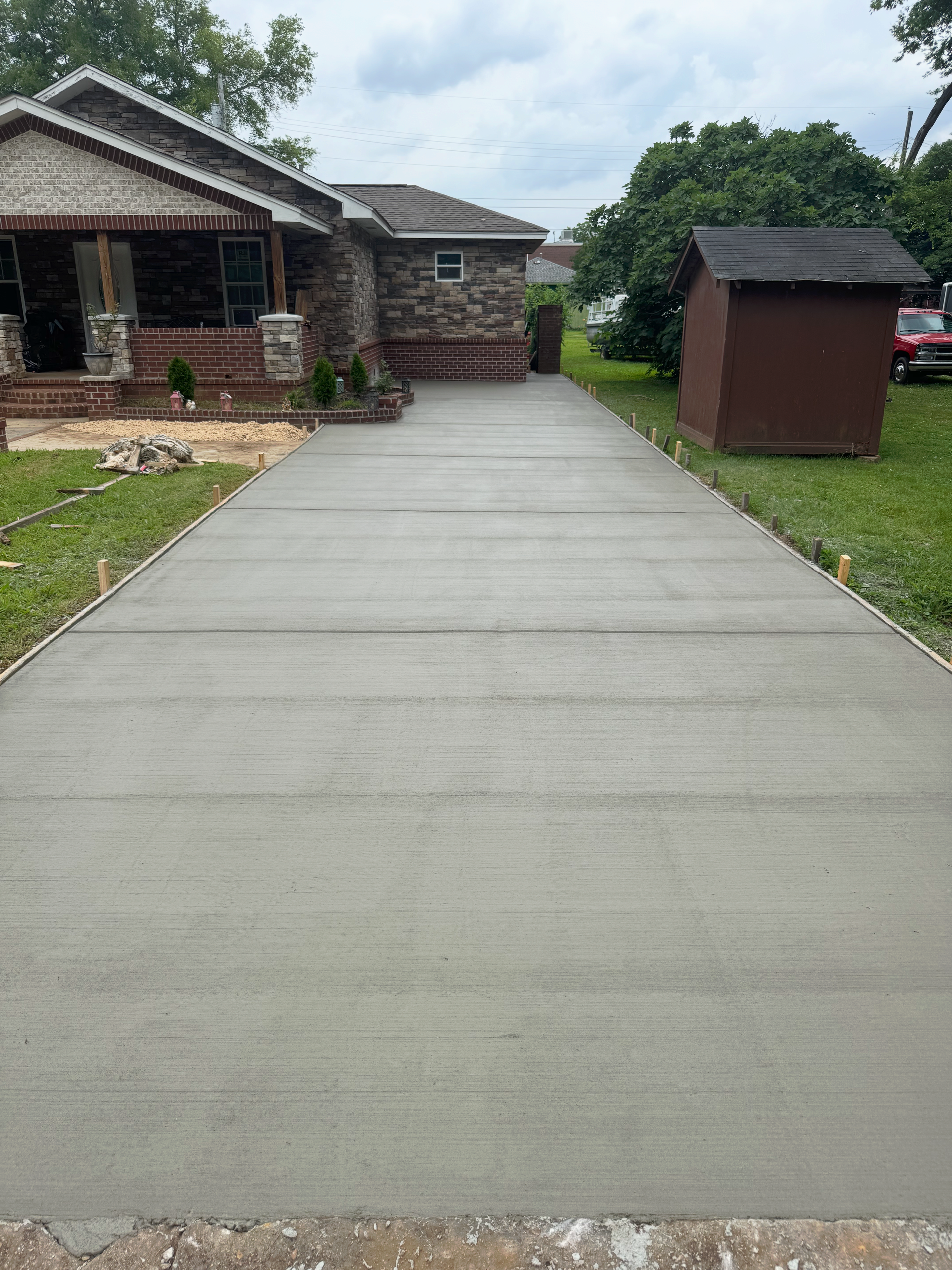 Concrete driveway leading to a brick house with a grassy yard and a small brown shed.