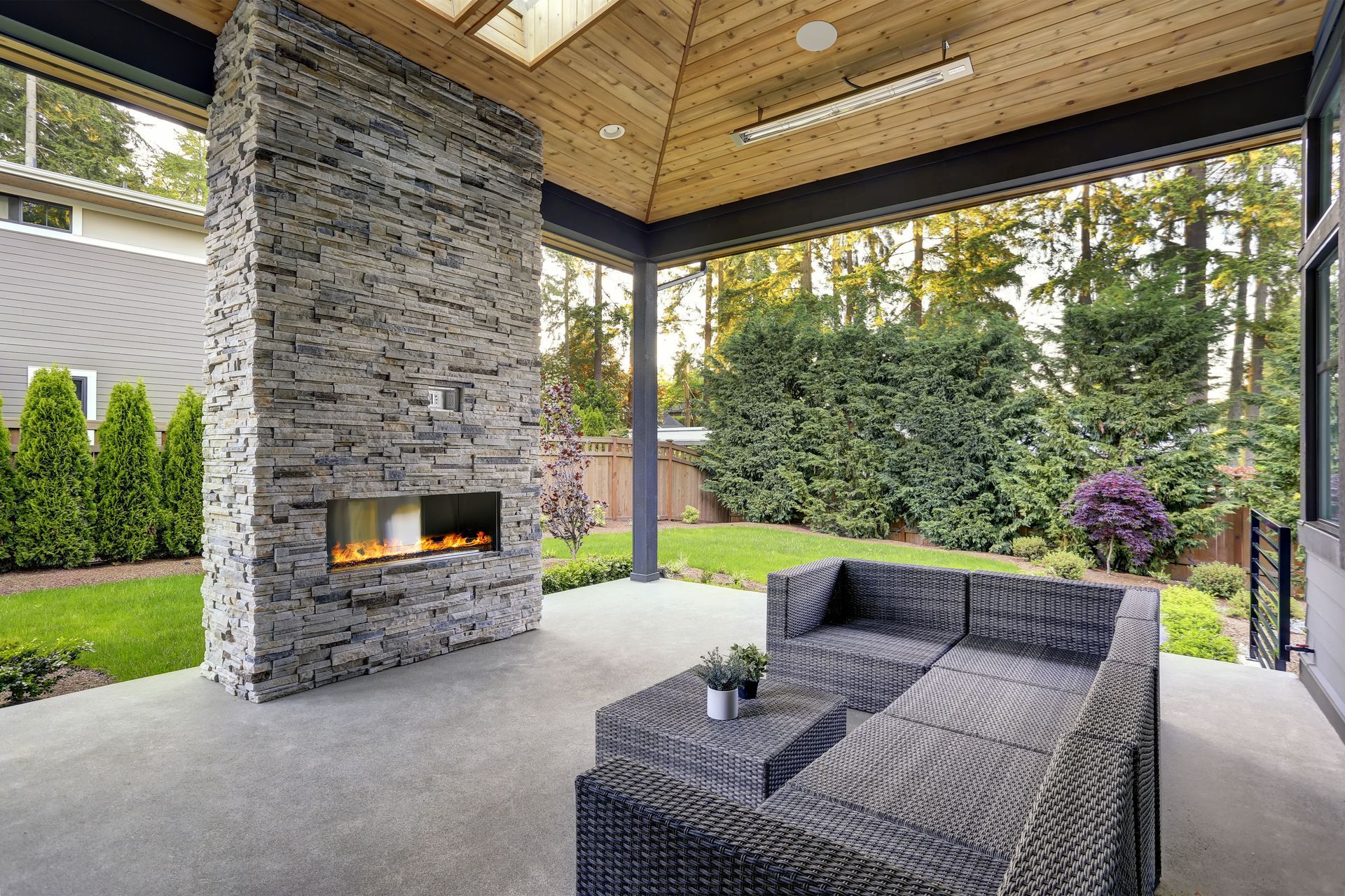 Covered outdoor patio with stone fireplace, gray wicker seating, and green backyard view