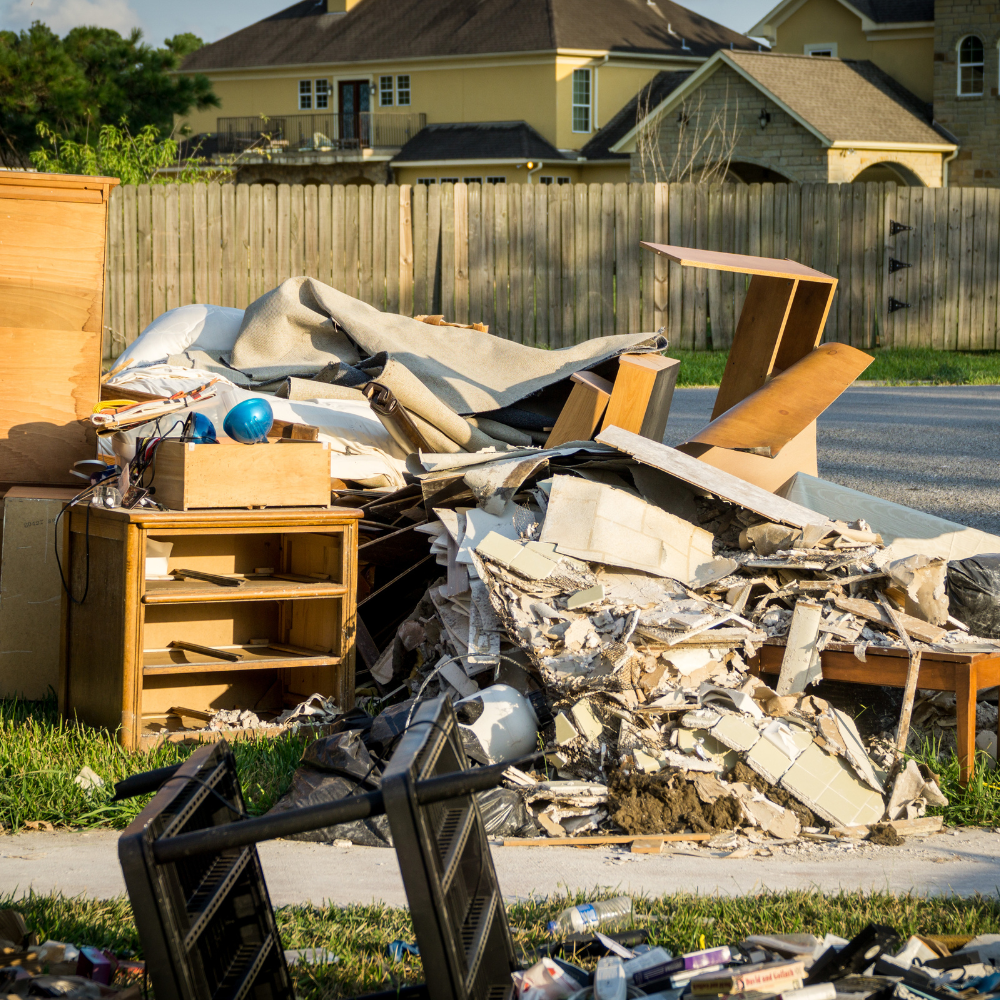 Pile of discarded furniture and debris on a curb in front of houses; wood, metal, beige and blue items visible.