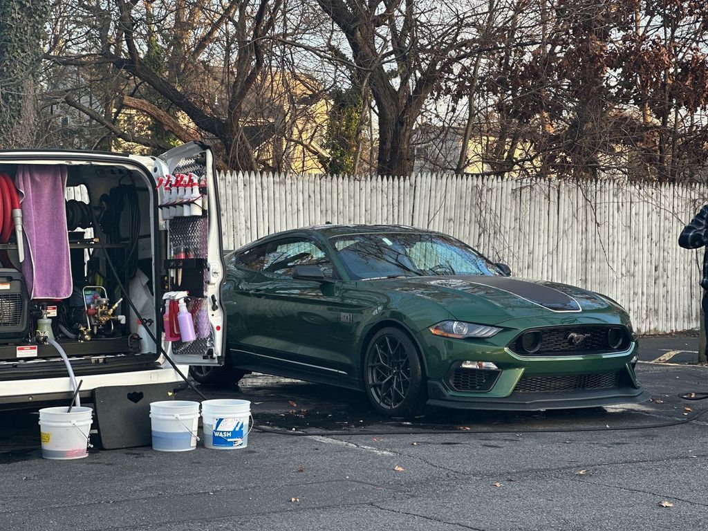 Green sports car parked beside an open trailer with gear in a wooded lot