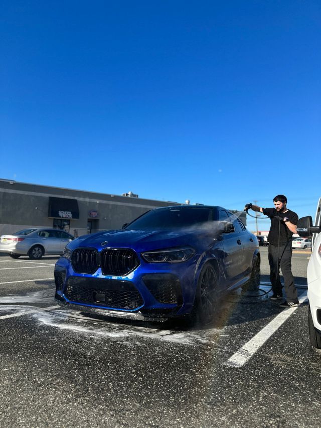 Blue BMW SUV being washed in a parking lot, with a person rinsing it under a bright blue sky.