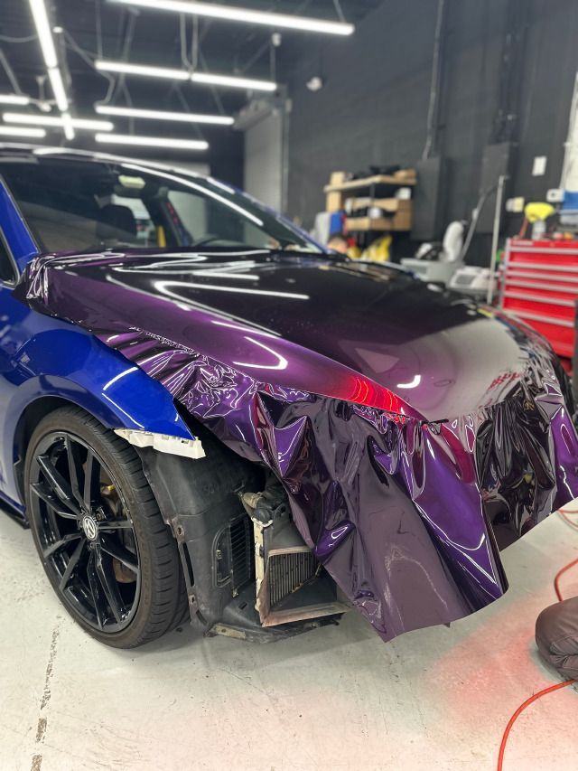 Purple and blue car in a repair shop, front end covered in wrinkled protective film