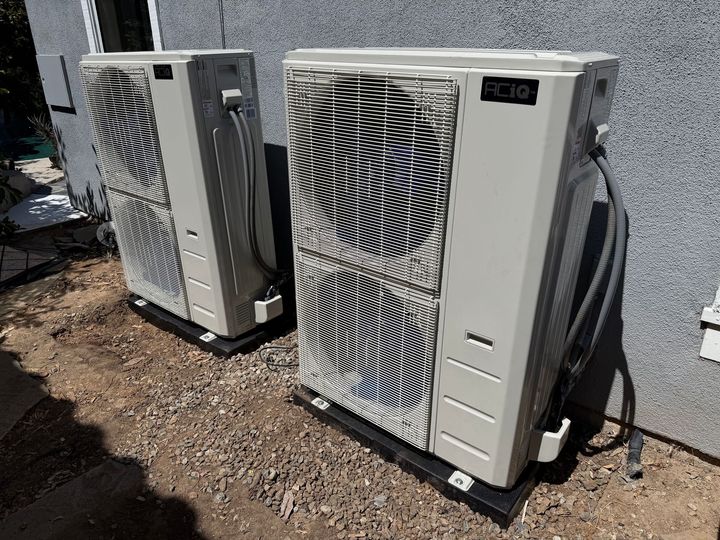 Two outdoor HVAC heat pump units beside a building on gravel, with coiled hoses and vents visible