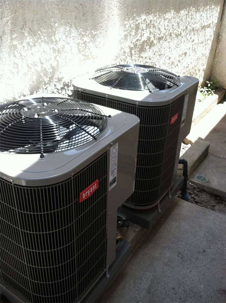 Two outdoor HVAC condenser units beside a stucco wall on a concrete pad.