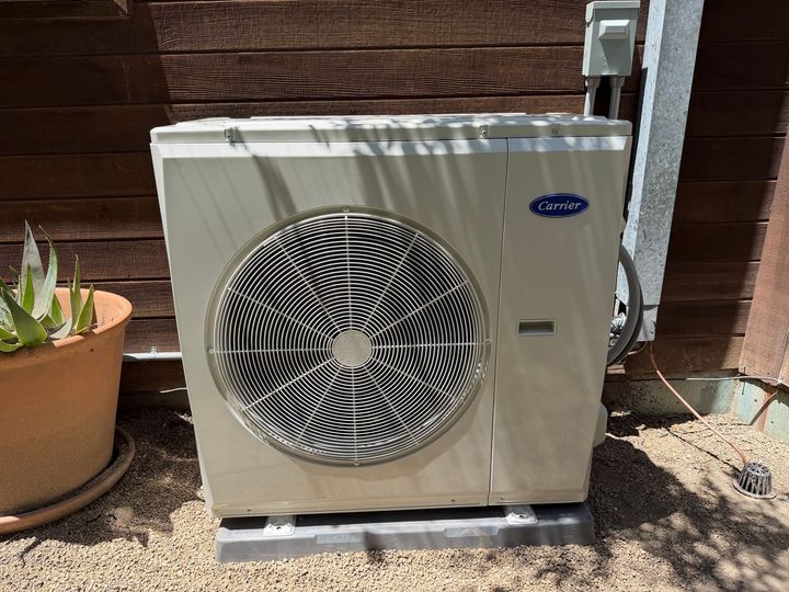 Outdoor air conditioner unit beside a potted plant against a wooden fence.