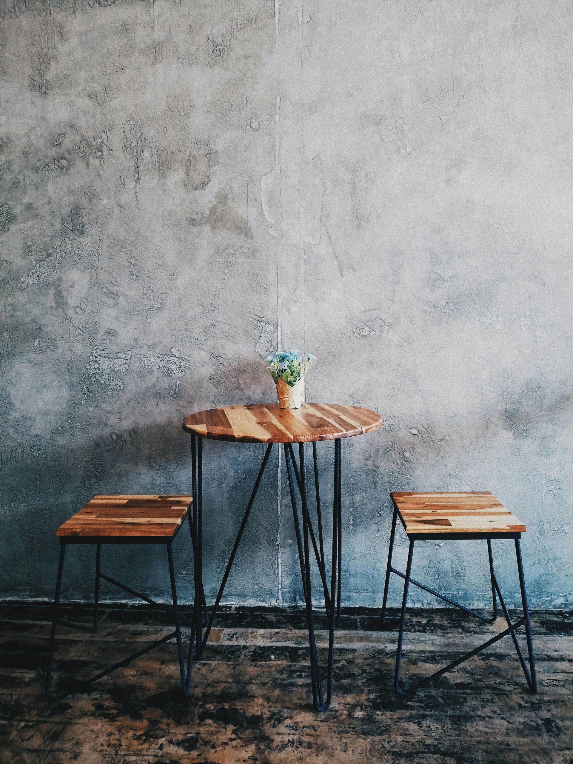 A wooden table and two wooden stools in front of a concrete wall.