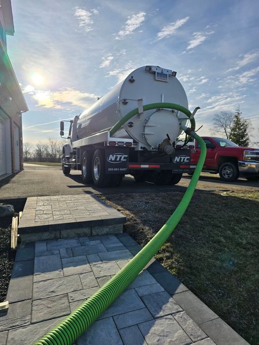 White tanker truck connected to a green hose on a sidewalk, parked beside a red pickup at sunrise.
