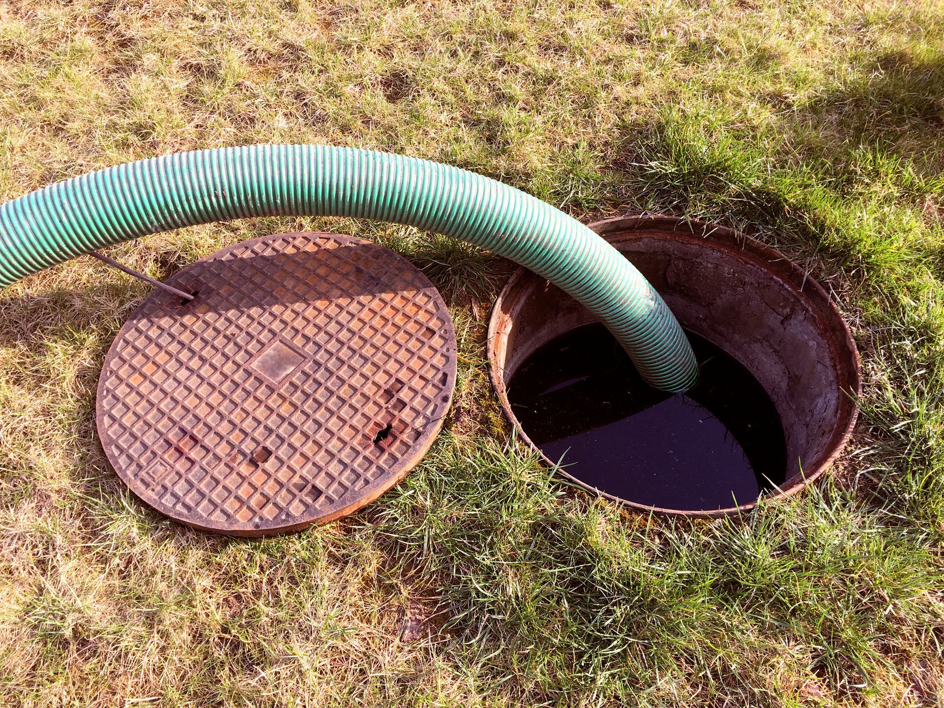 Green hose draining into open manhole on grass, with the round cover beside it