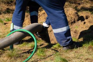 Worker in blue pants using a hose near a dirt trench in a grassy field