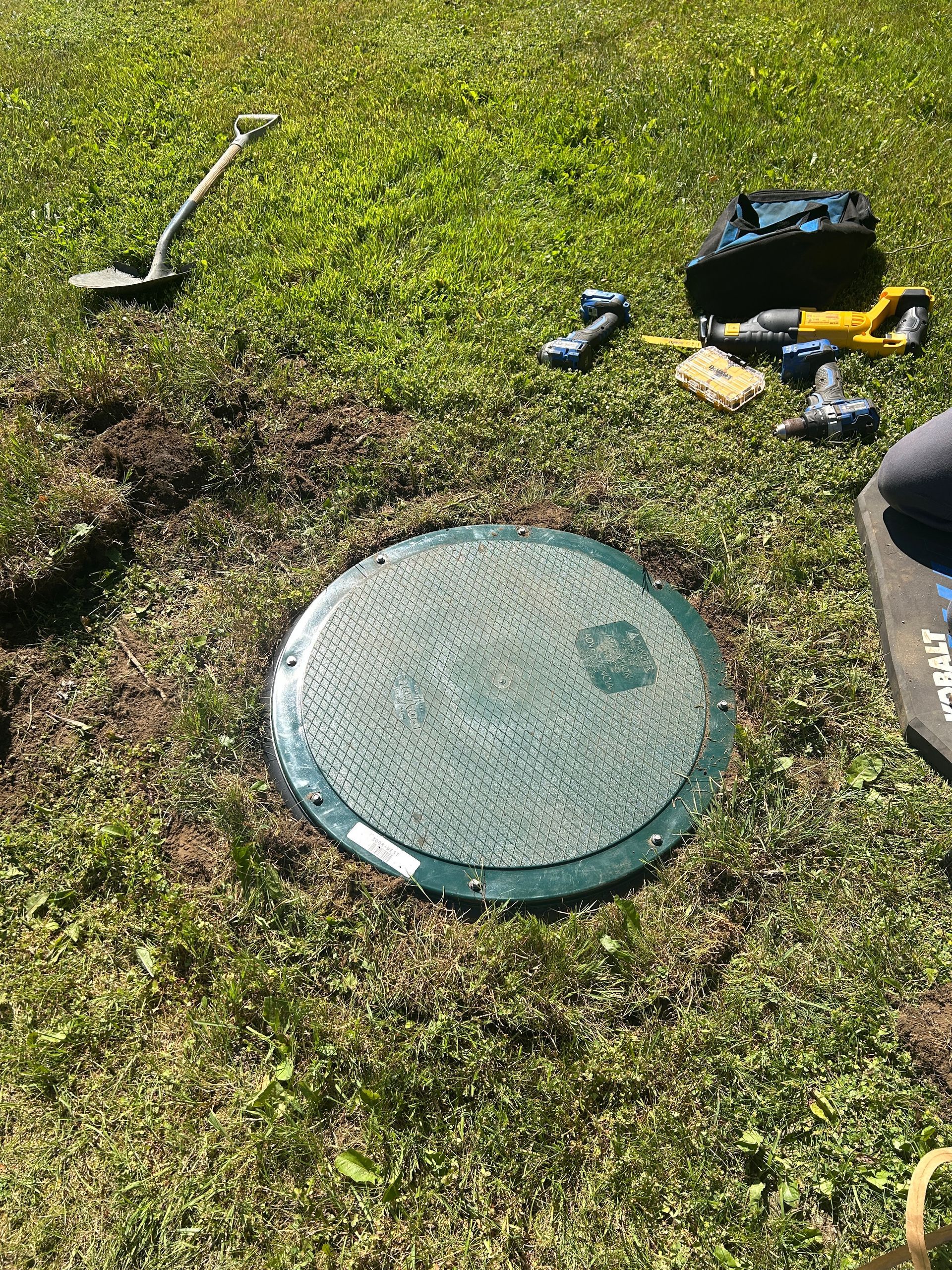 Round black manhole cover set in dirt beside grass and rocks