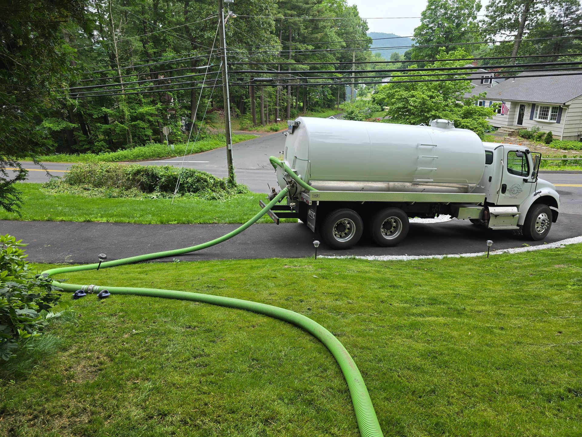 Green hose connected to white tanker truck on a curved road by a grassy, wooded area