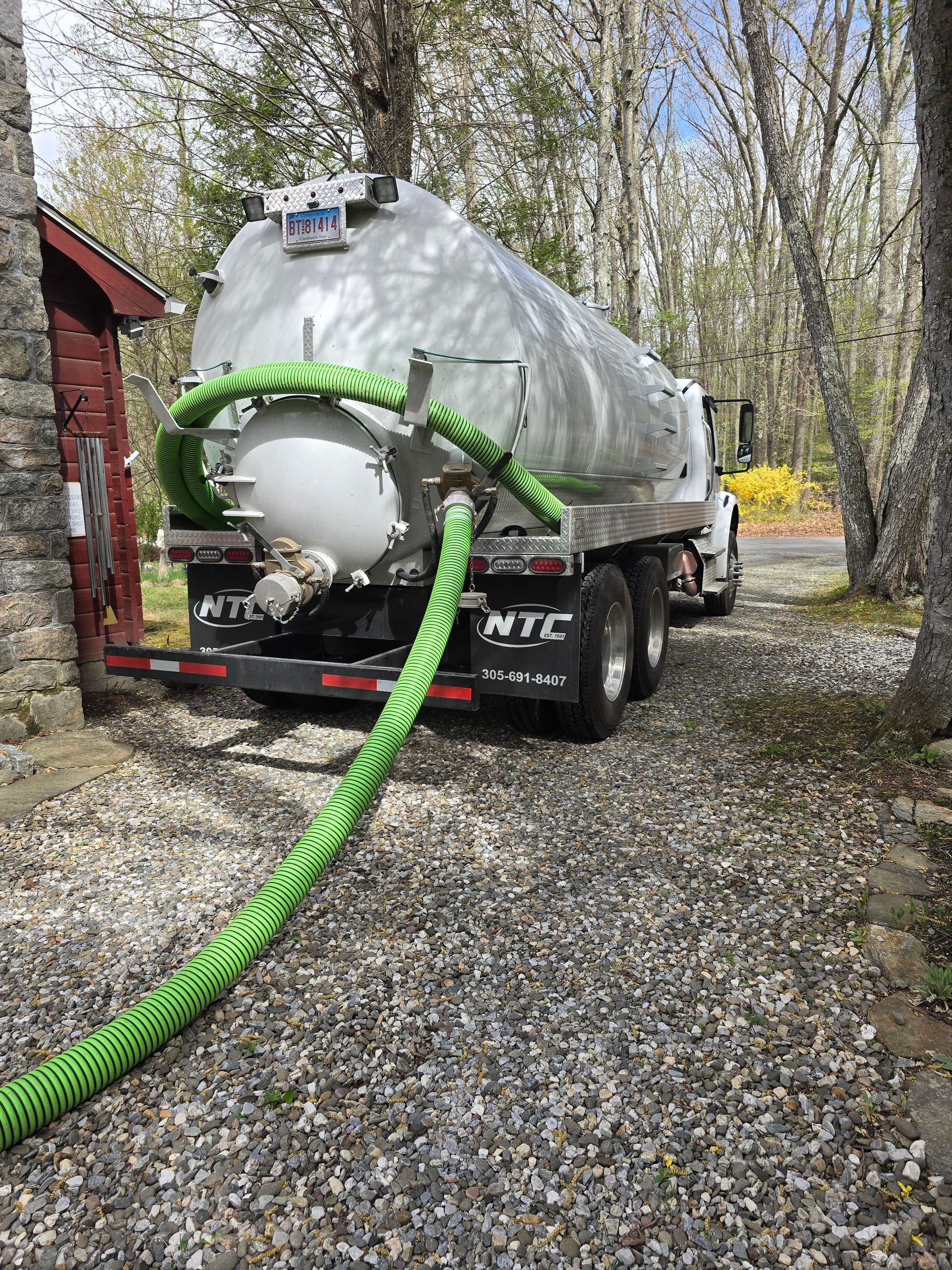Silver tanker truck with green hose parked on gravel beside a red building in a wooded area
