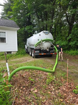 White hose or pipe runs across a dry yard from a metal tank to a small house.