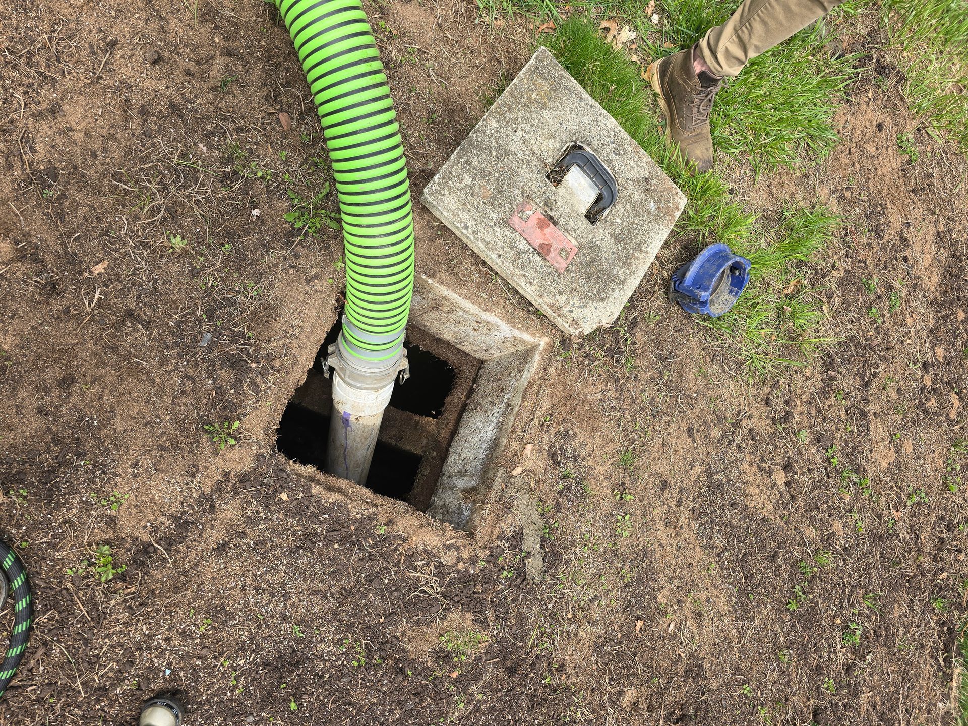 Green hose enters an open utility hole beside a square concrete cover on bare ground.