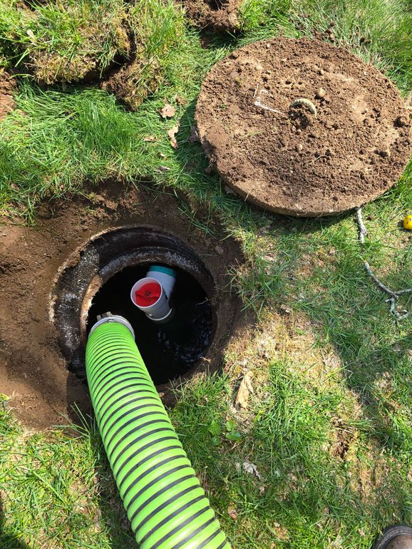 Green corrugated hose in a ground access hole beside a round dirt cover on grass