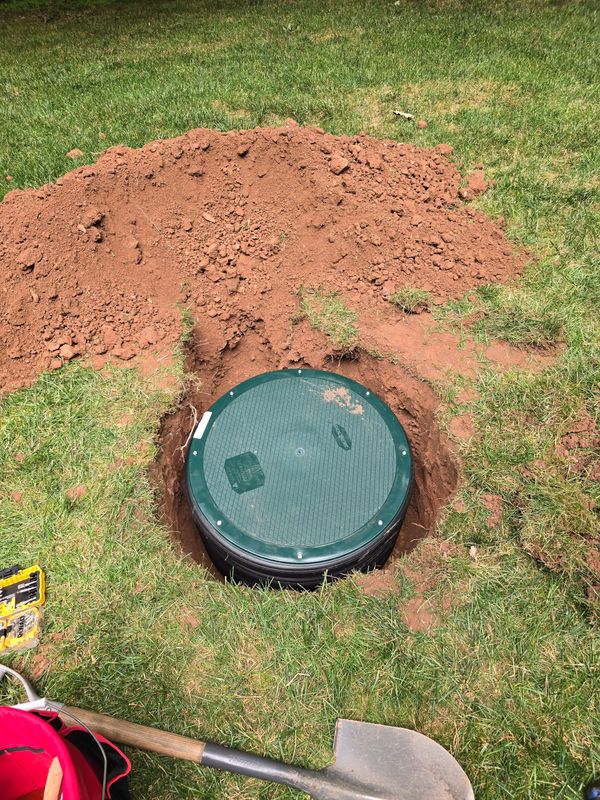 Green circular manhole cover in a grassy yard beside a pile of reddish dirt and a shovel handle.