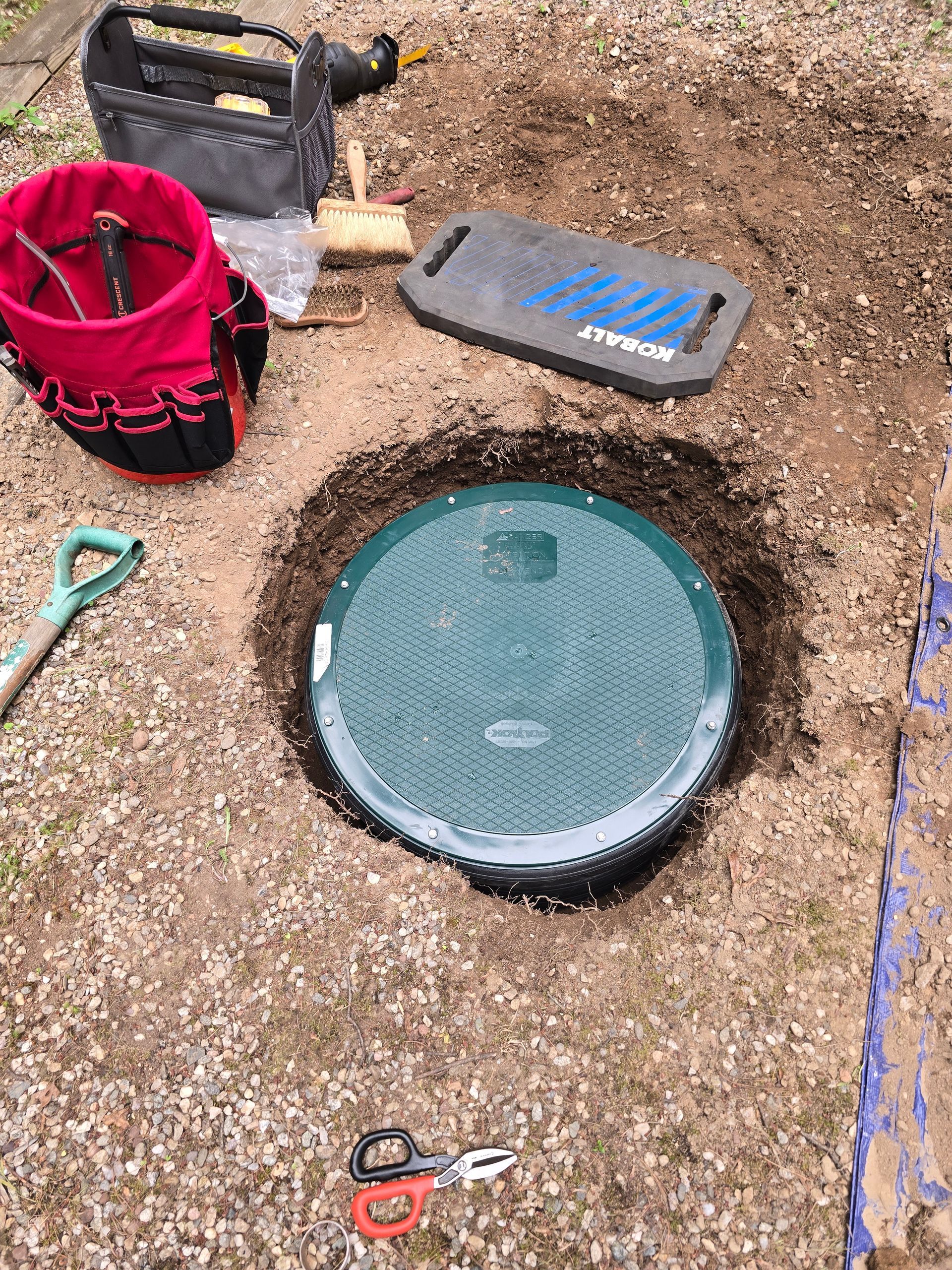 Round pool drain cover in dirt with tools and a red bucket nearby outdoors