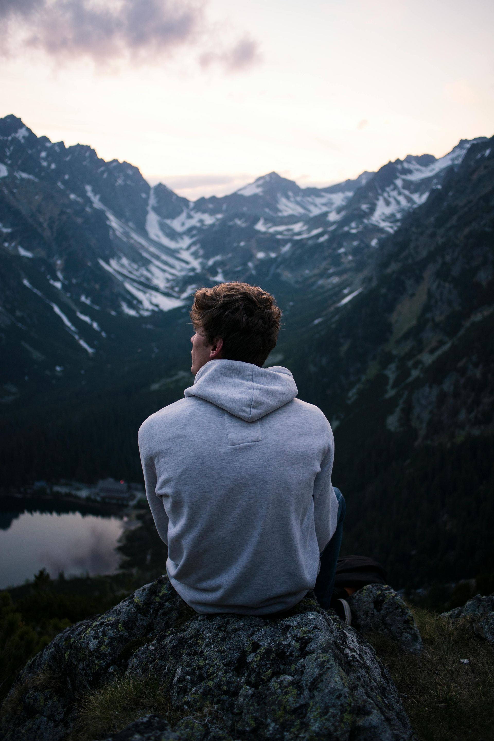 Person in a gray hoodie sits on a rock overlooking a mountain valley at dusk.