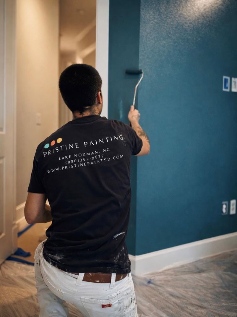 Painter rolling blue paint on an indoor wall in a room with white trim.