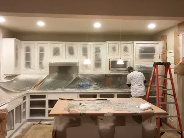 Kitchen being painted white, with cabinets masked off and a worker on a red ladder.