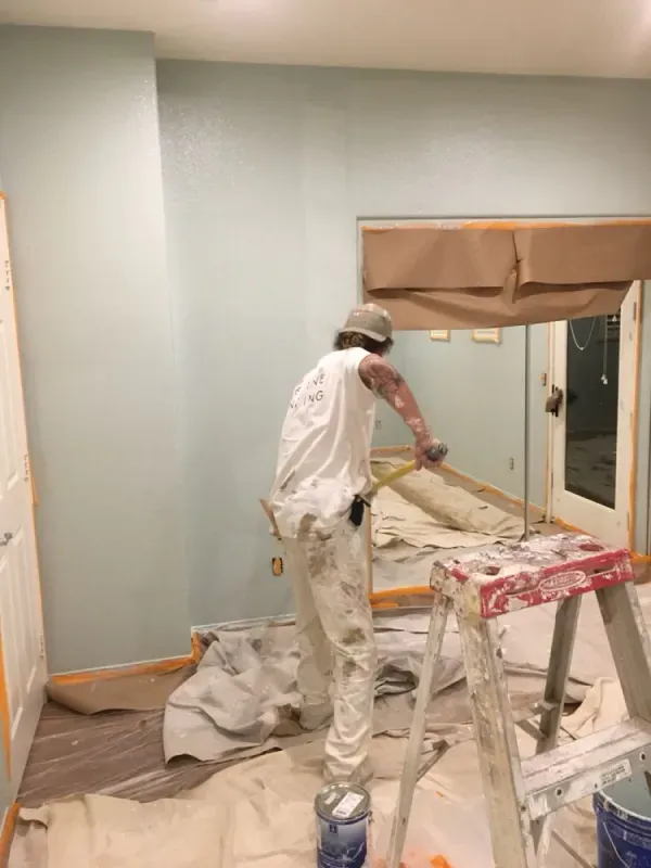 Person painting a room with white walls and a tan window shade, using a roller amid drop cloths and supplies