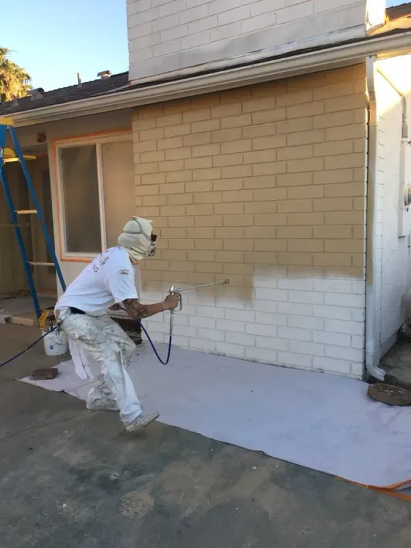 Person spray-painting a brick wall white beside a house exterior