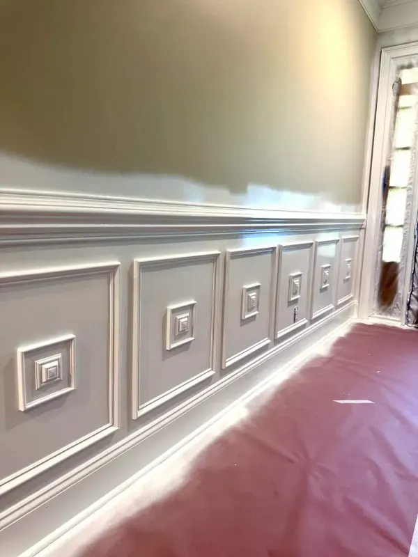 White paneled hallway wall with chair rail molding and a red floor runner, lit by daylight from a doorway