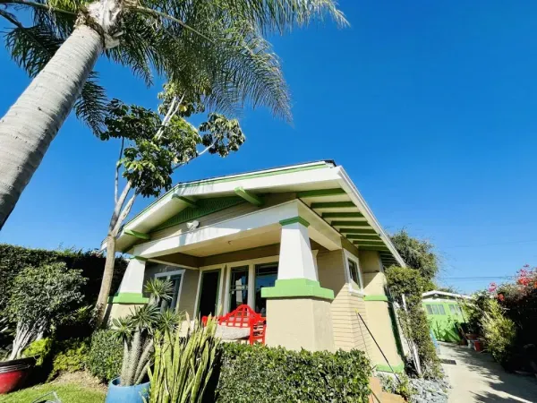 Small tropical house with green trim, red chairs on the porch, and palm trees under a bright blue sky
