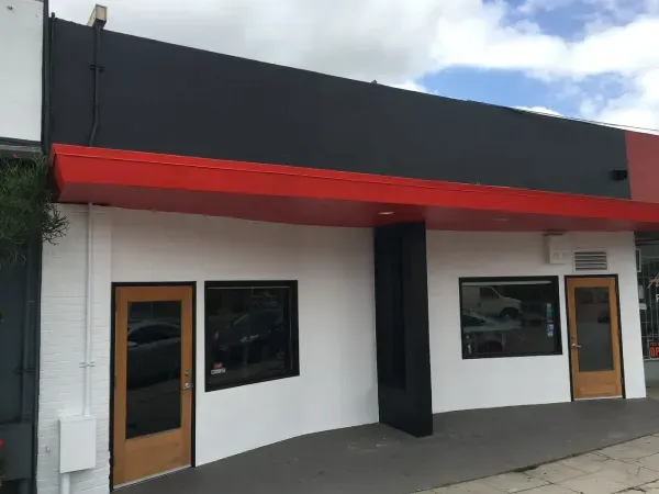 Small storefront with white walls, black trim, red awning, and two wooden-framed windows and doors