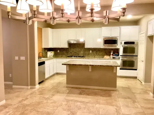 Modern kitchen with white cabinets, granite island, and large chandelier over beige tile flooring