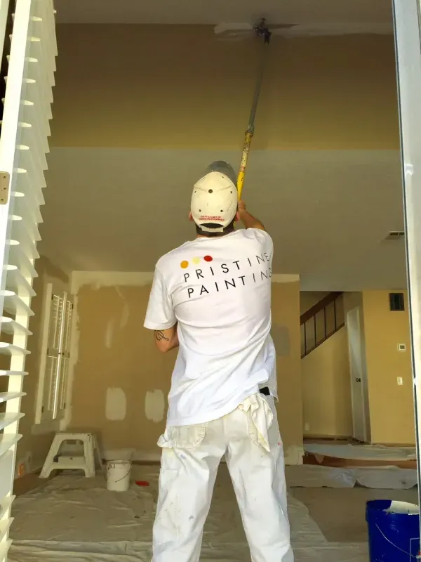 Painter roller-coating a beige room, wearing white clothes and a mask on a step stool.