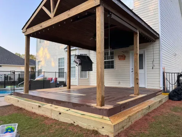 Covered wooden patio deck beside a white house, with a sloped roof and railings.