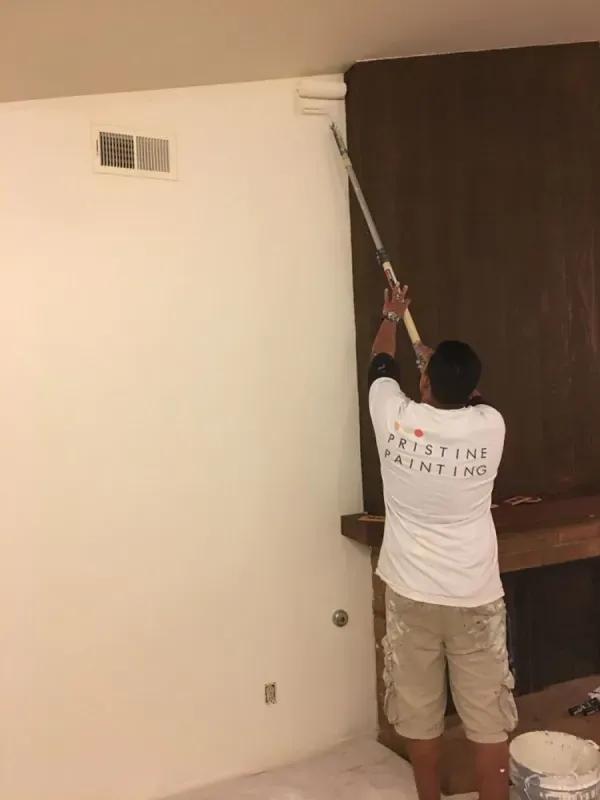 Person painting a ceiling corner with a long roller in a room with white walls and dark wood paneling