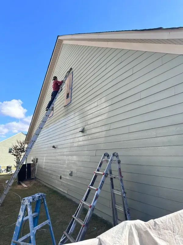 Worker painting exterior siding on a house with ladders set against the wall.