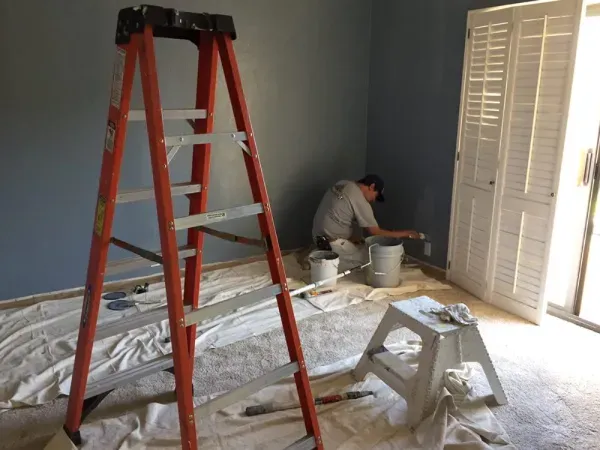Home renovation scene with red ladder, plastic-covered floor, and worker painting near a bright window