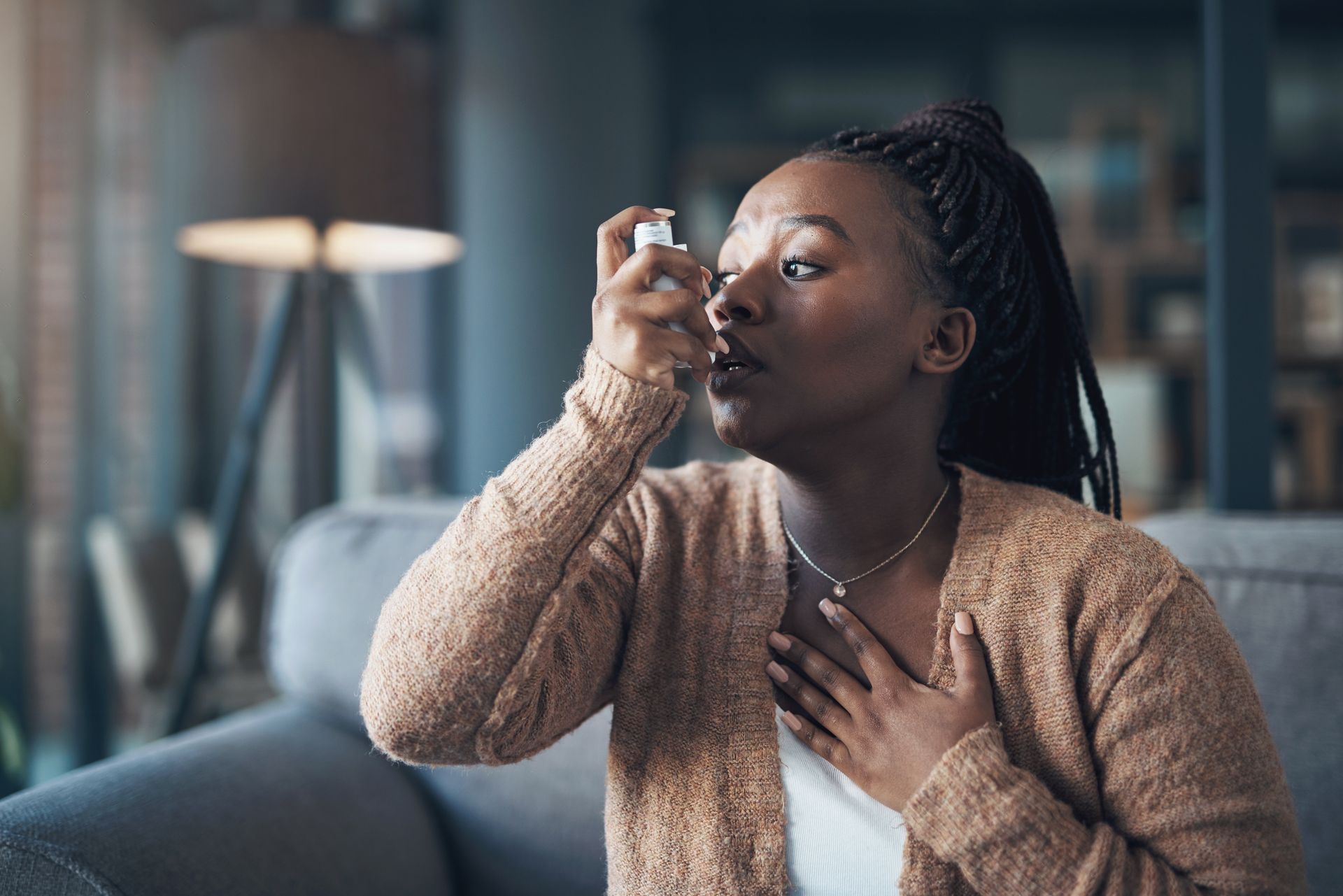 Woman using an inhaler indoors, with a hand on her chest, in a living room.