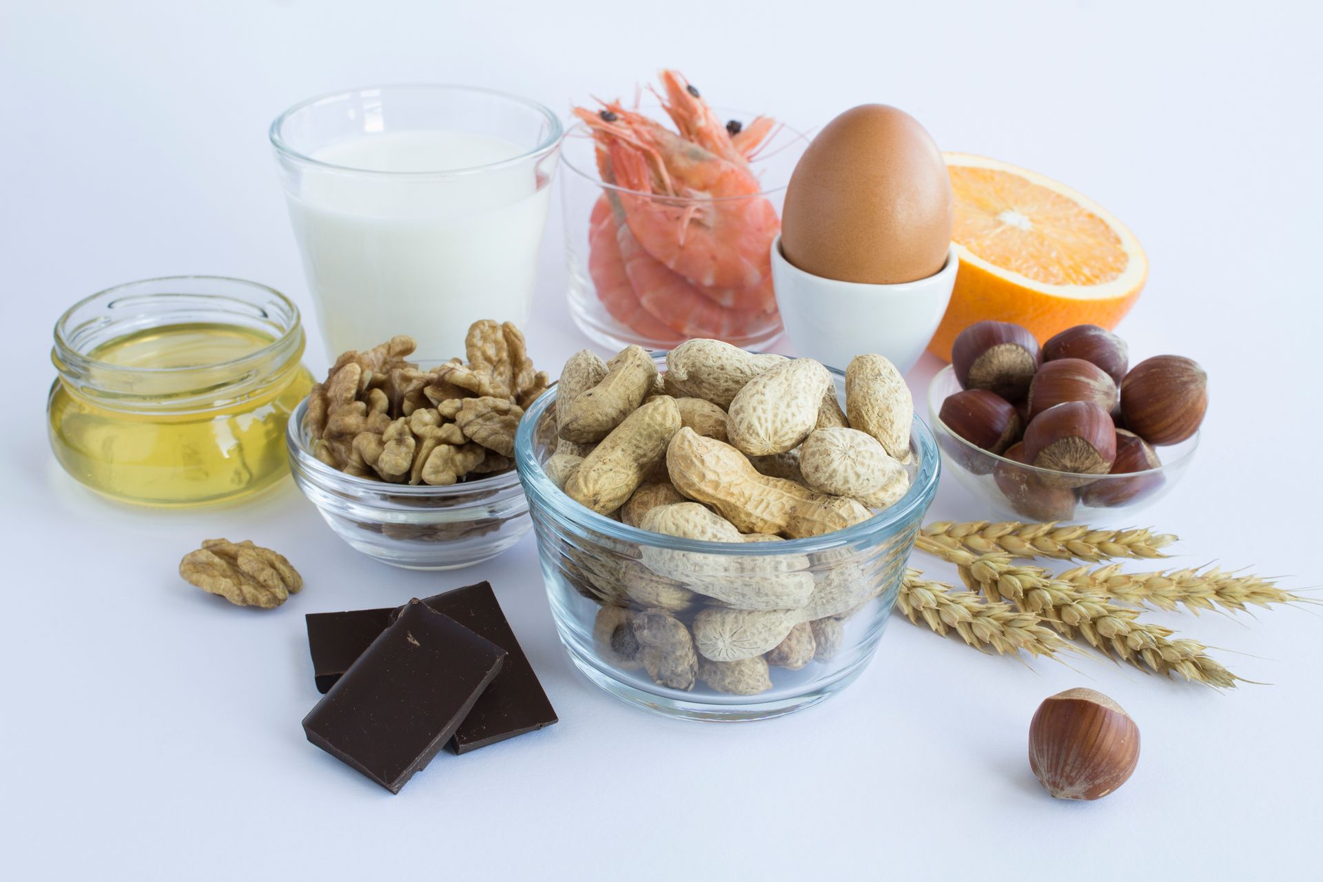 Assortment of healthy foods on a white surface: milk, eggs, nuts, shrimp, orange, grains, chocolate, and oil.