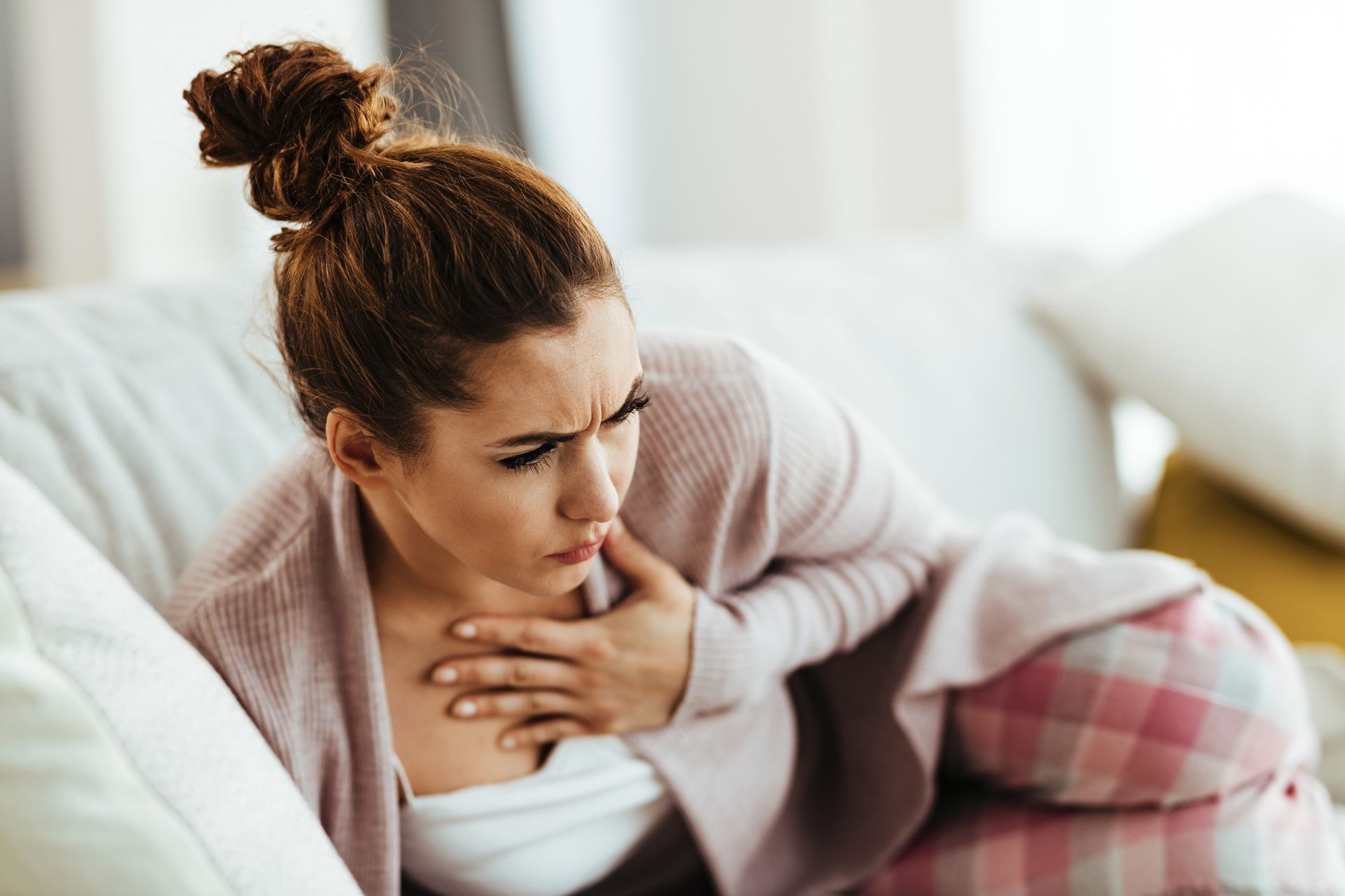 Woman sitting on a couch, clutching her chest and looking uncomfortable in a bright living room