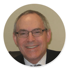 Smiling man wearing glasses in a suit and tie against a plain beige background