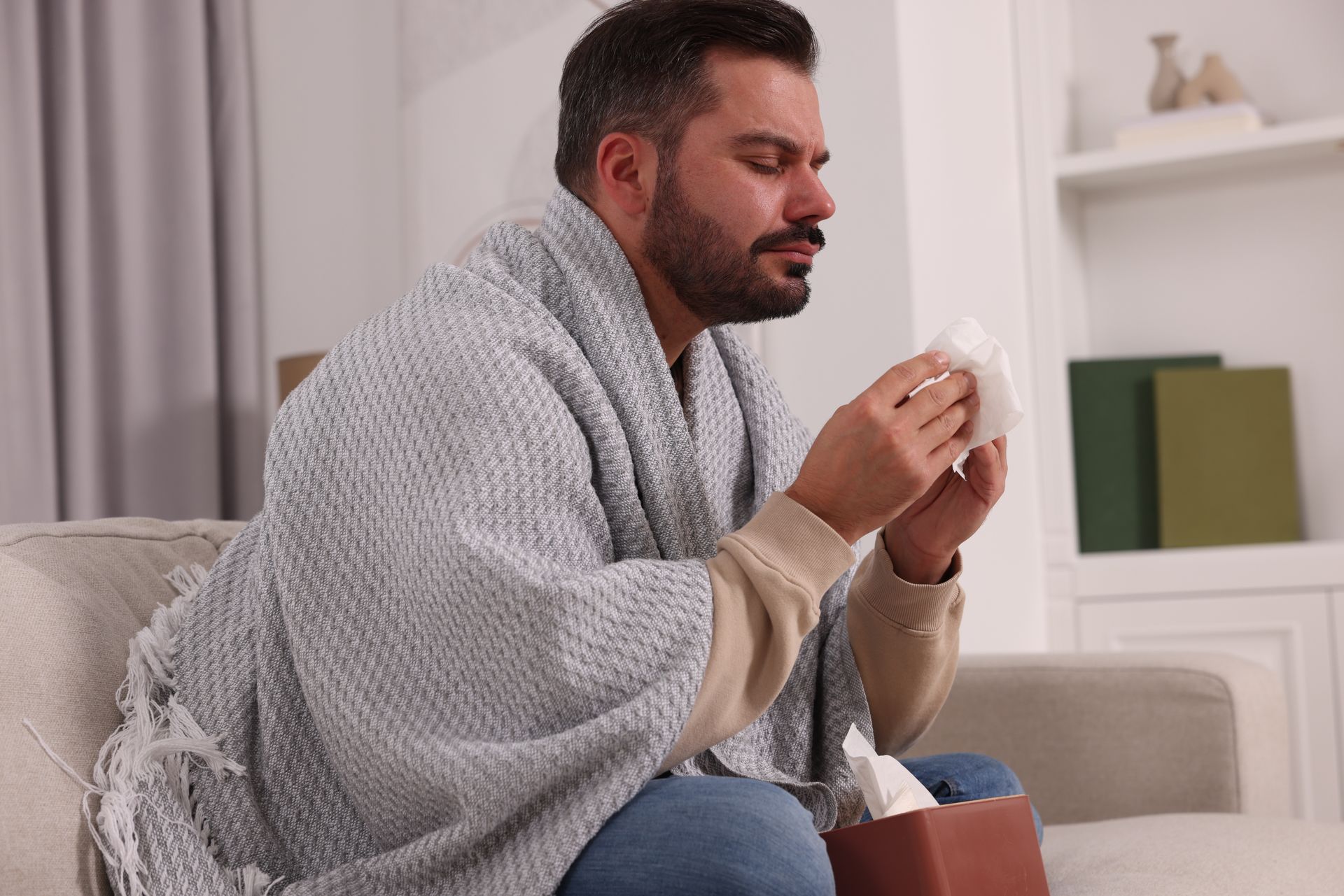 Man sitting on a couch wrapped in a gray blanket, holding a cup in a cozy living room.