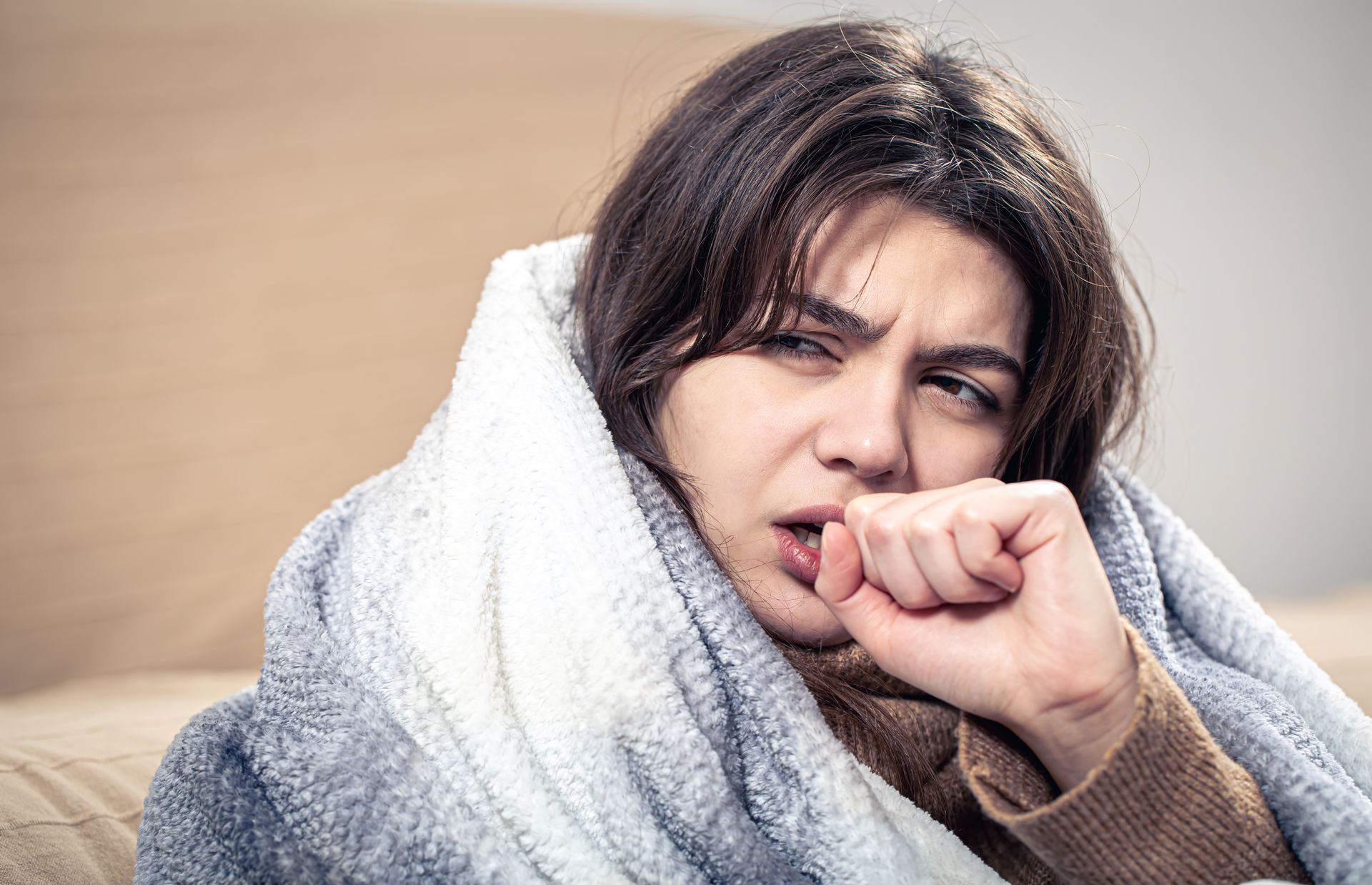 Woman wrapped in a white blanket coughing into her fist, looking unwell outdoors
