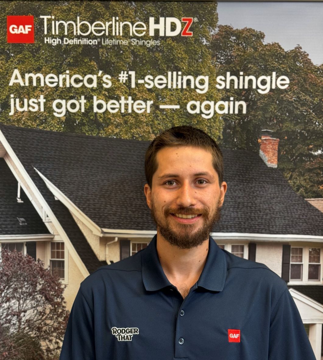 Man in navy polo standing before a GAF Timberline HDZ shingle advertisement backdrop