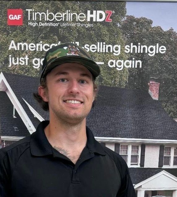 Smiling man in black shirt and cap standing before a GAF Timberline HDZ roofing advertisement